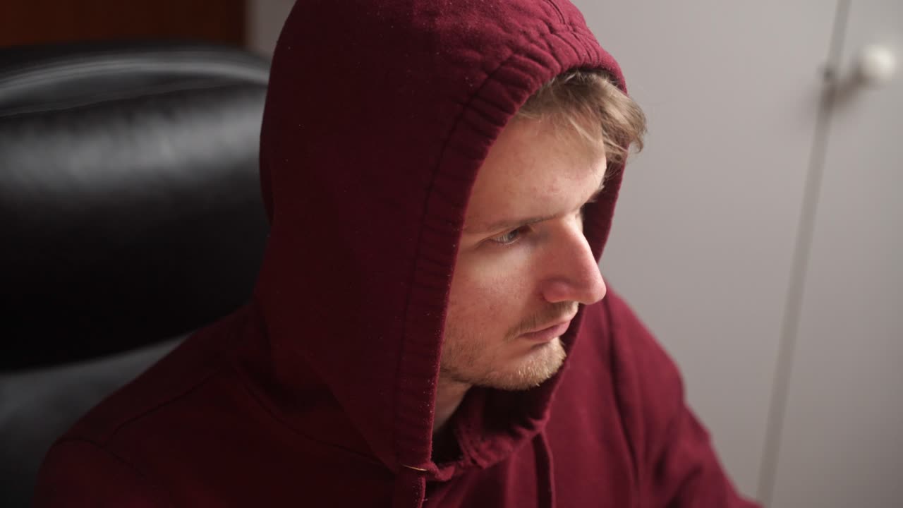 un joven con una sudadera con capucha de la universidad se sienta en una silla de oficina en casa y procede a ejecutar un código en una computadora