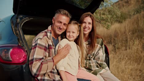 Portrait-of-a-happy-family-who,-during-their-picnic-outside-the-city,-sits-in-the-open-trunk-of-a-black-car-and-poses.-A-happy-brunette-girl,-together-with-her-husband,-the-middle-aged-man-with-gray-hair-in-a-checkered-shirt,-and-their-little-daughter,-the-blonde-girl,-are-sitting-in-the-open-trunk-