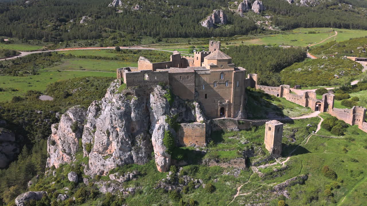 Backward drone shot rising to reveal a medieval castle atop a rocky cliff, surrounded by green hills, dense forest, and ancient defensive walls under a clear spring sky