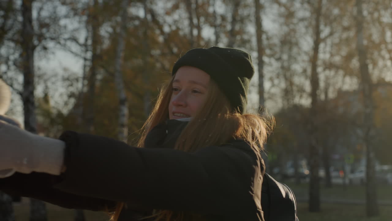 Женщина протягивает руку, держа игрушку, в городском парке на закате. Задумчивое выражение лица, видны шарф и рюкзак. Медленное движение с мягким контровым светом. Меланхоличное настроение, намекающее на прощание или воспоминания, кинематографический стиль.