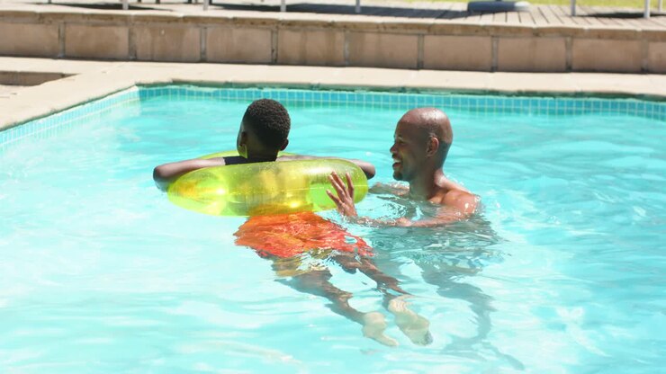 African man supporting boy in orange trunks and ring, guiding learning, boy kicking at home pool