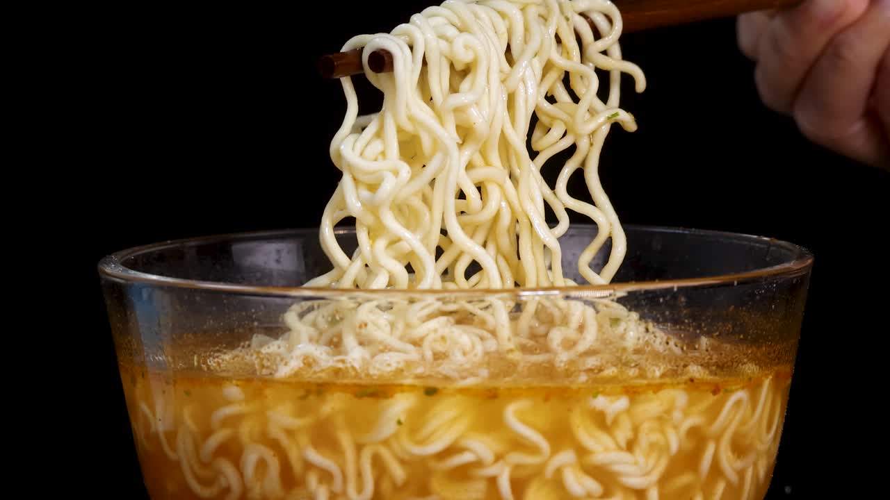 Hand uses chopsticks to lift cooked instant noodles from a clear glass bowl filled with broth, set against a black background with even lighting