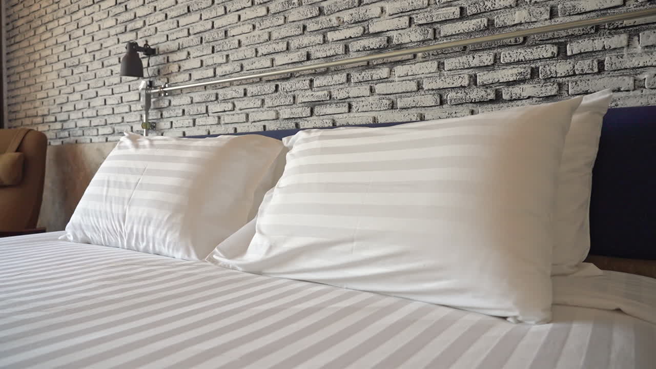 Slow pan right on hotel made-up bed, striped pillows on brick wall background