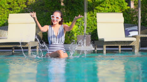 una mujer bonita sentada entre dos tumbonas en la parte poco profunda de la piscina mientras salpica agua de la piscina en el aire