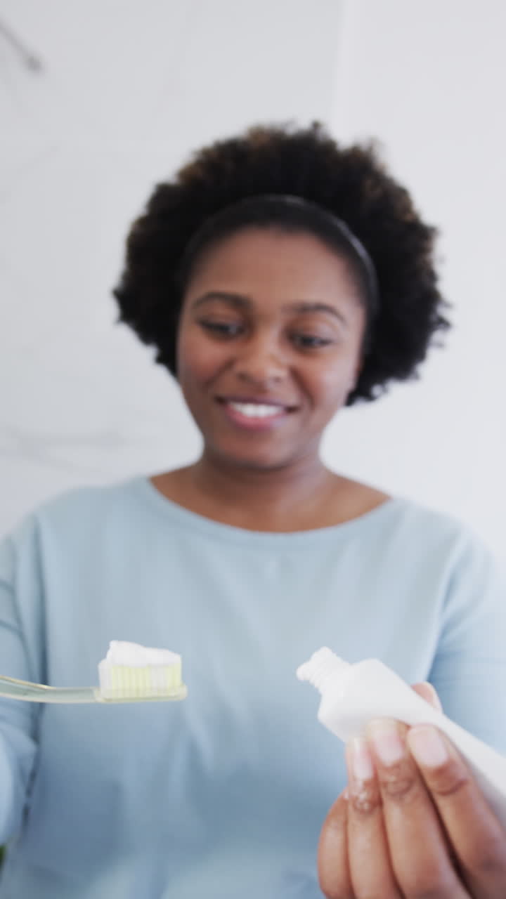 Vertical video of portrait of african american woman brushing her teeth on light background