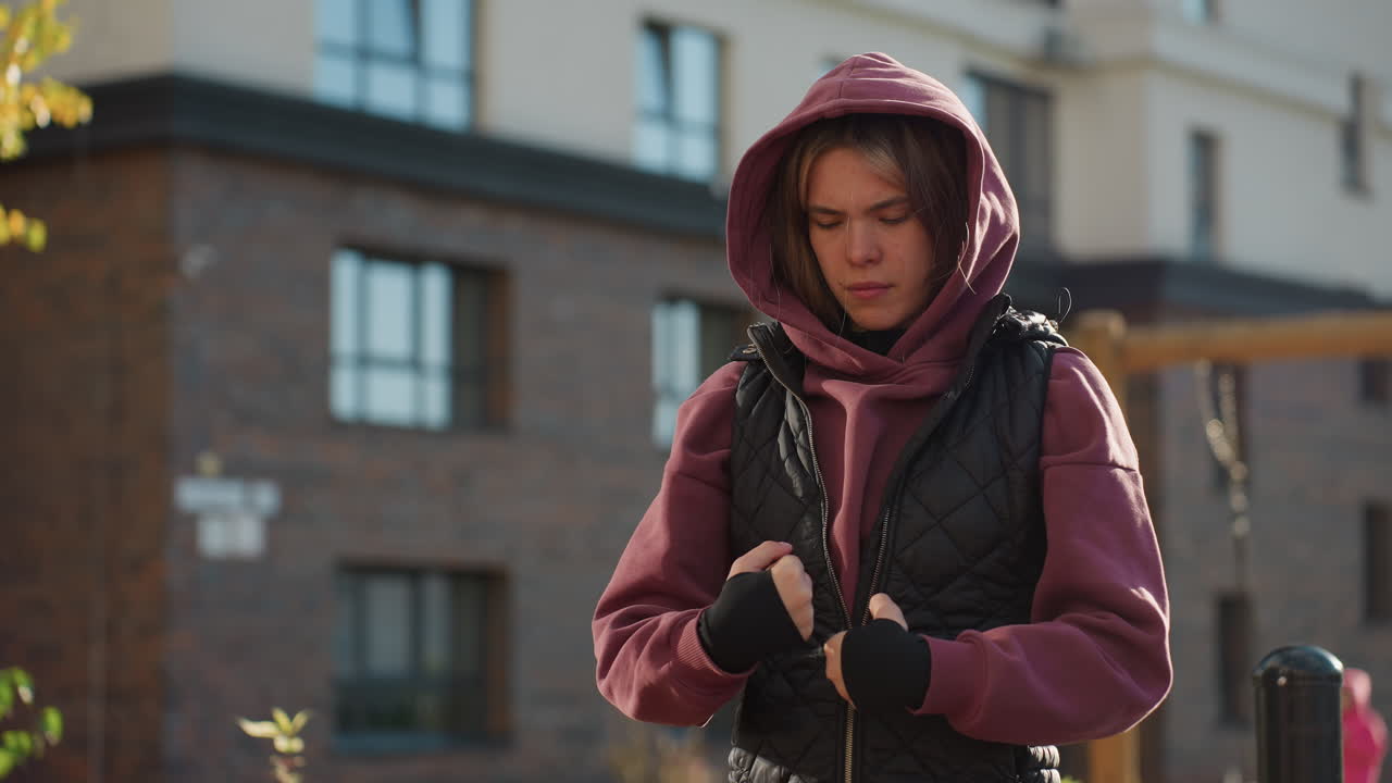Balanced performer wearing hoodie and black gloves rolls hands gently with focused expression while warming up outdoors in urban autumn setting with sunlight highlighting movement for fitness