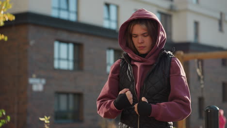 balanced performer wearing hoodie and black gloves rolls hands gently with focused expression while warming up outdoors in urban autumn setting with sunlight highlighting movement for fitness