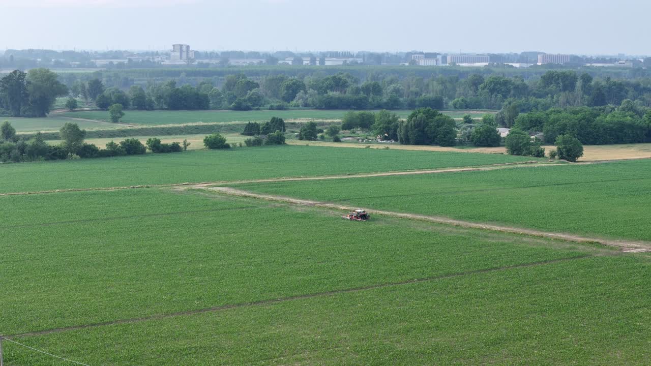 Aerial shot of tractor with boom sprayer operating in green maize field, with Po River and Lombardy skyline visible in the background, illustrating modern intensive tecniques in growing season