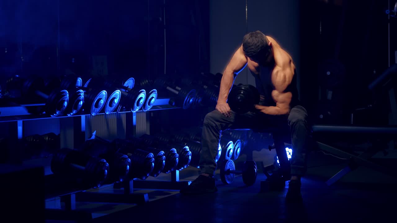 Muscular man working out in gym doing exercises with dumbbells, strong male naked torso, abs. Blue light filter in sport gym. Selective focus.