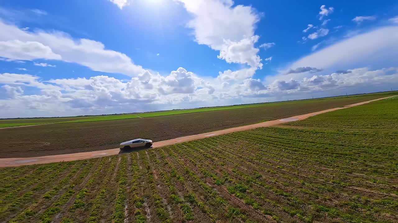 Ride of a futuristic Tesla Cybertruck by the farmlands. FPV drone footage of a modern auto with a blue sky at backdrop.