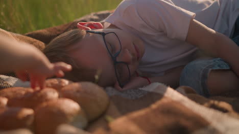 a close-up of a boy wearing glasses and a white shirt, lying peacefully on a checkered scarf in a grassy field. next to him, a fresh loaf of bread rests, adding a cozy, serene atmosphere