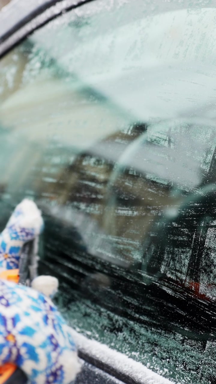 Close-up of gloved driver scraping frost and snow from a car side window with a windshield scraper, restoring visibility on a cold winter morning. Vertical b-roll.