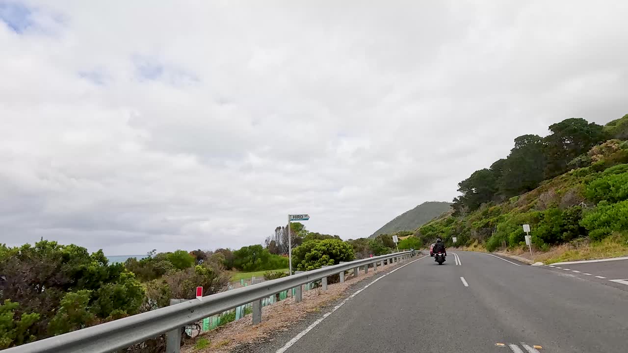Motorcyclists ride along the scenic Great Ocean Road with lush greenery and ocean views under cloudy skies