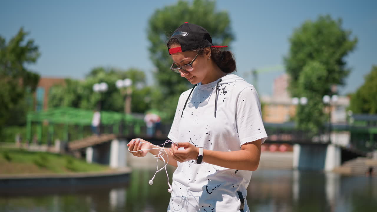 young woman counting beads by lake, practicing quiet mindfulness with hands moving gently over string while sunlight shimmers on water green trees, bridge backdrop, casual summer outfit, meditative