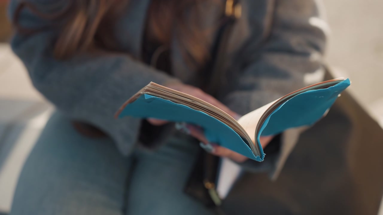 Pěkný tyrkysový zápisník v rukou studentky kavkazského vzhledu sedící na lavičce v šedém kabátě, listující stránkami a dělající si poznámky perem. Detailní záběr na texturovaný kožený obal, šité vazby a okraje papíru.
