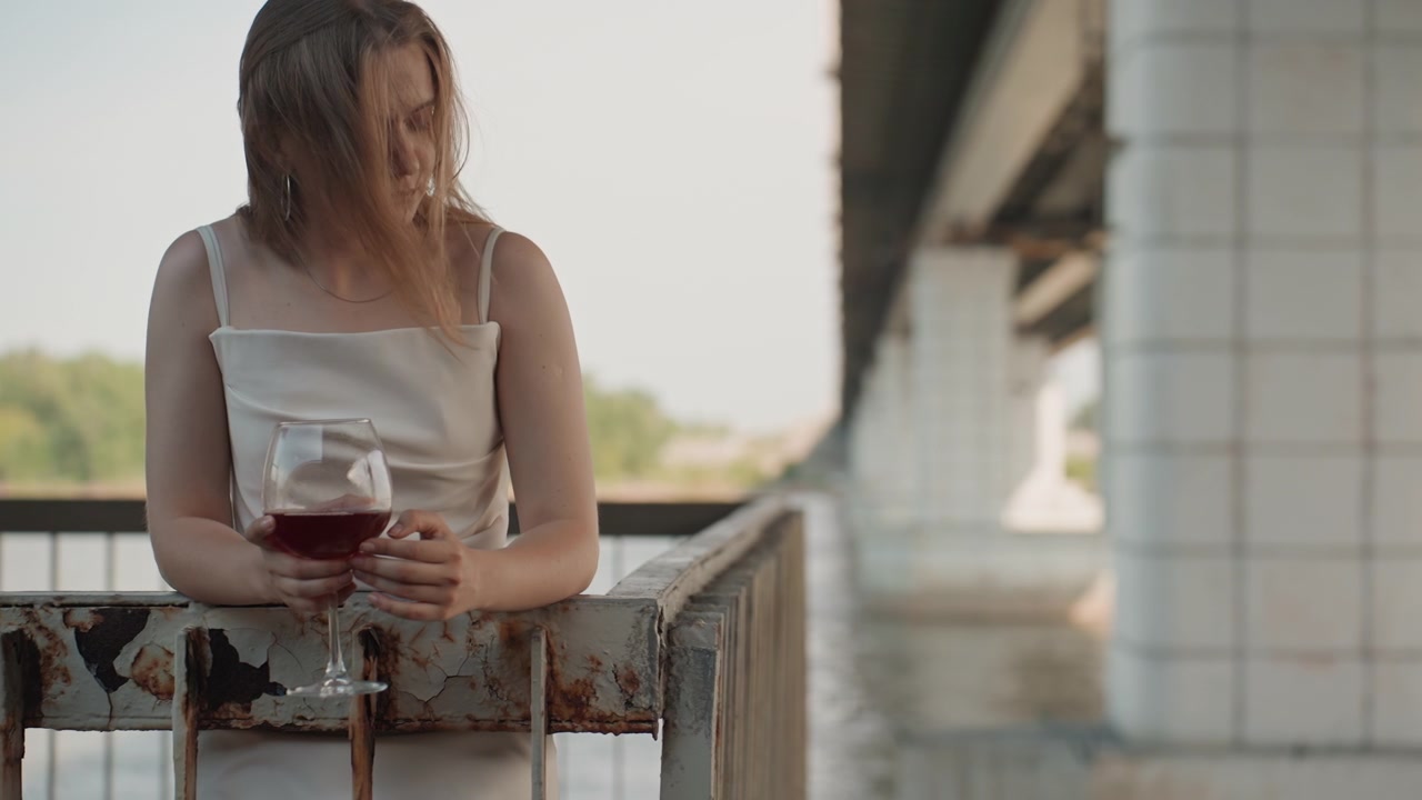 Una donna in abito bianco è in piedi su un ponte, tiene e osserva un bicchiere di vino rosso. La scena è caratterizzata da una luce naturale soffusa con il vento che le scompiglia i capelli mentre la donna si appoggia a ringhiere metalliche usurate.