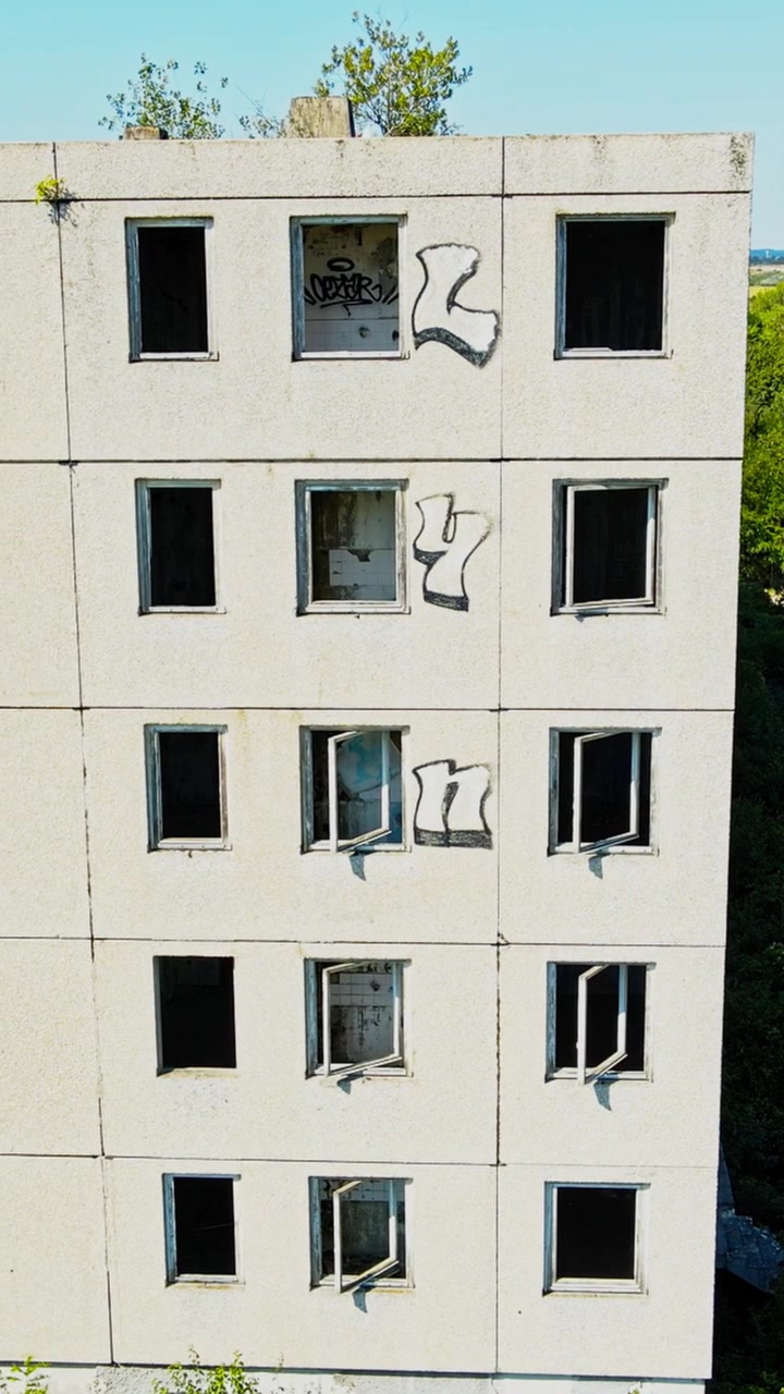 Video vertical con imágenes aéreas que rodea un bloque de hormigón desierto y marcado con grafitis, con ventanas rotas, invadido por densa vegetación verde bajo la luz del día brillante. Perfecto para historias de redes sociales