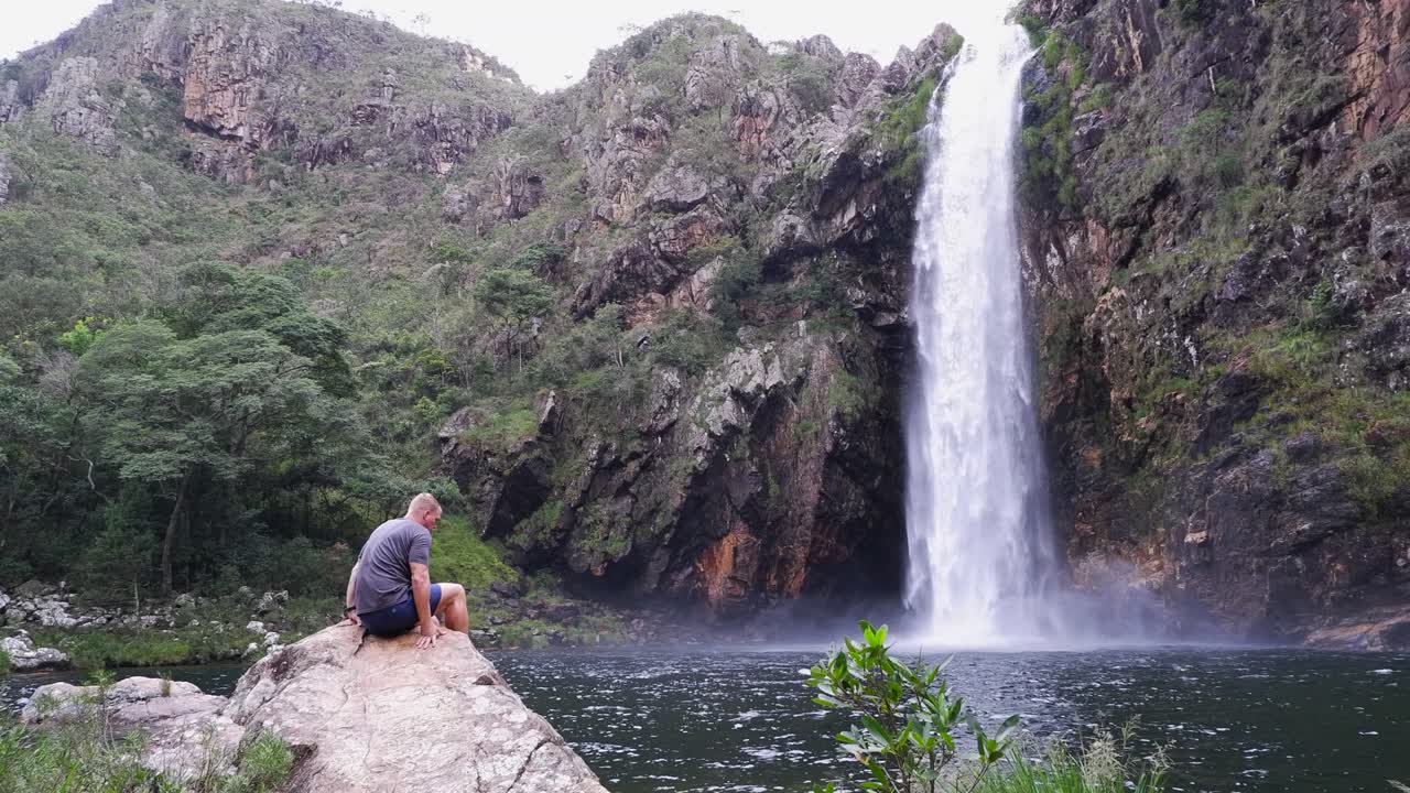 Young man sits on boulder at misty pool below Fundao Waterfall, Brazil