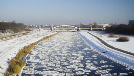 Gefrorener-Fluss-Mit-Eisschollen-Mit-Einer-Brücke-Und-Der-Stadt-Im-Hintergrund