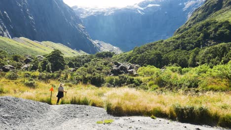 female hiker with hiking sticks walking downhill between rocks and grasses between mountains during bright sunny day