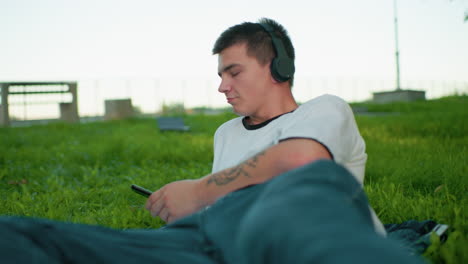 music artist enjoys listening to music outdoors, lying on his side with his hand propping up his head in a grassy field, background features an unclear object and a person walking in the distance