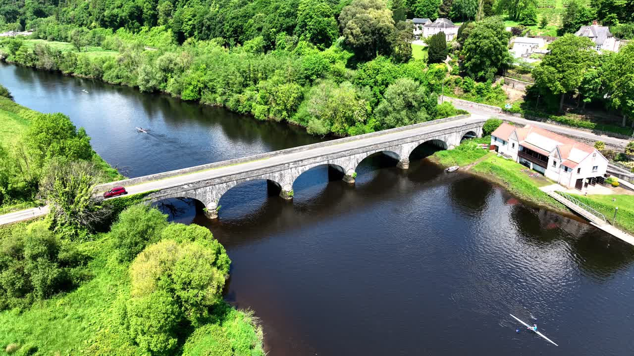 Aerial view of a river with an arch bridge, a red car, and rowers