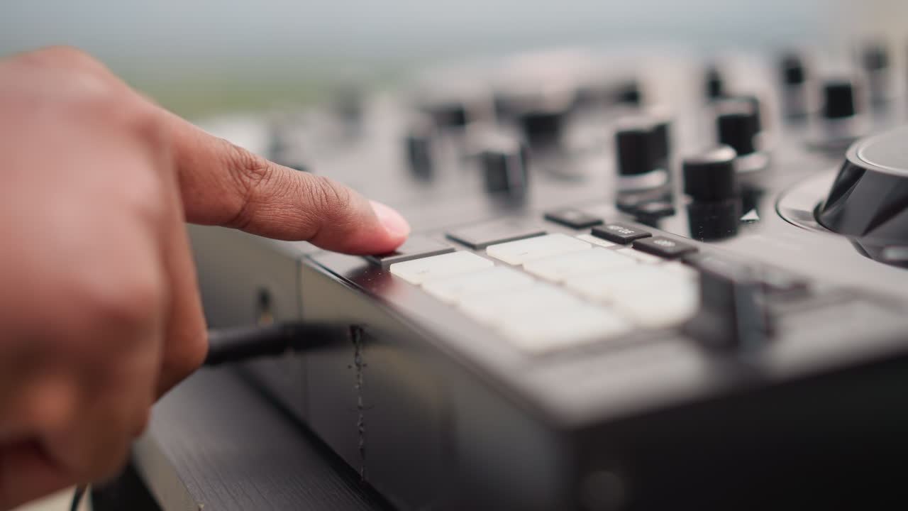 Tangan hitam dari dekat mengetuk pad drum pada kontroler kompak, tombol dan fader buram, kabel terpasang, pengaturan luar ruangan di siang hari, DJ jalanan yang fokus melakukan sampling loop lofi, ketukan jari yang presisi, minimalis.
