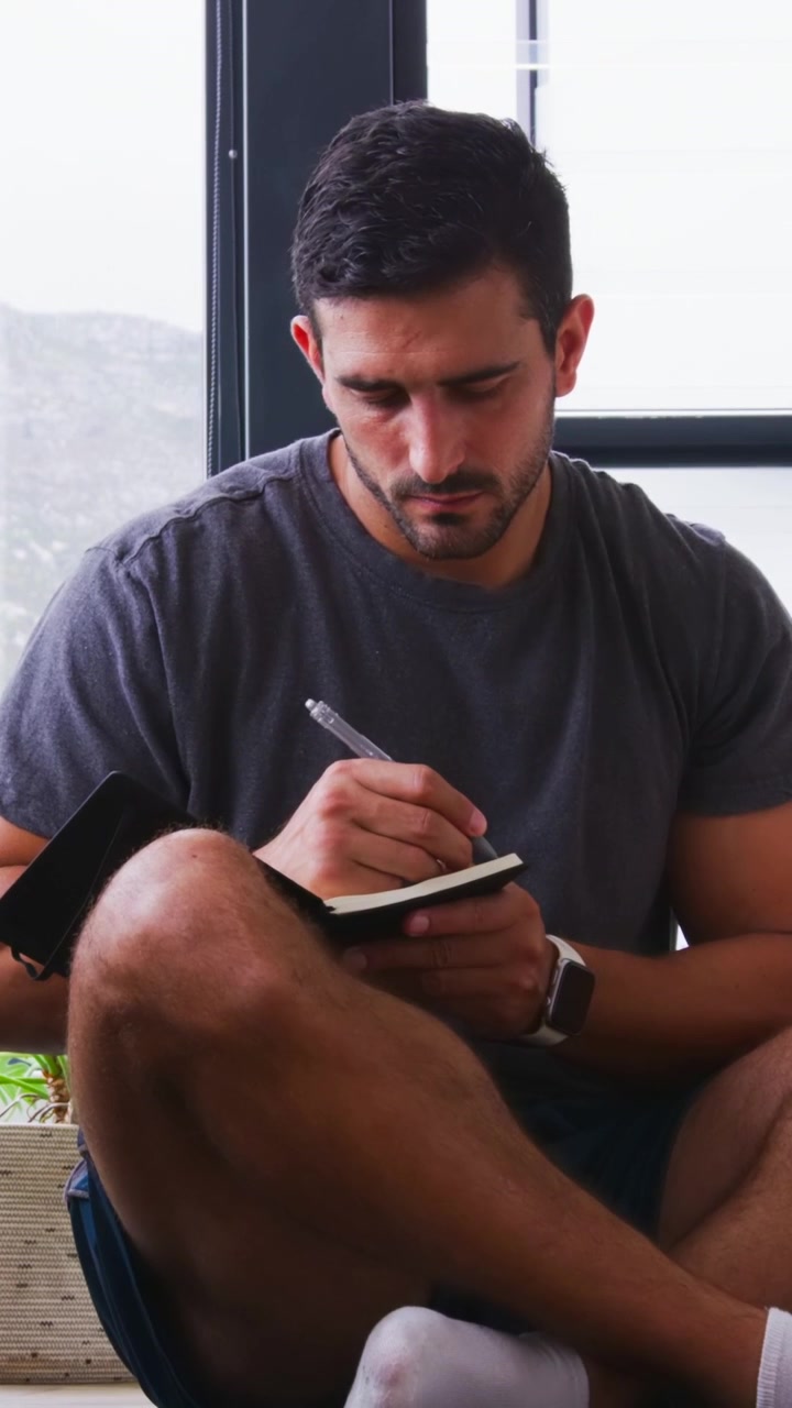 Vertical video: Opening black notebook, man taking notes on knee at home near window with pen