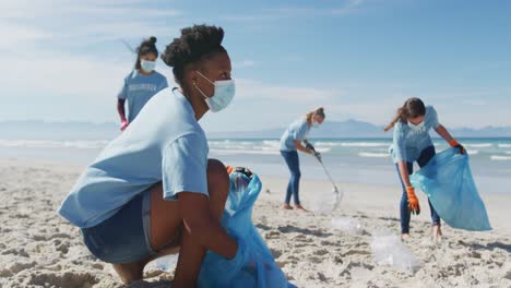 Diverse-group-of-women-wearing-volunteer-t-shirts-and-face-masks-picking-up-rubbish-from-beach