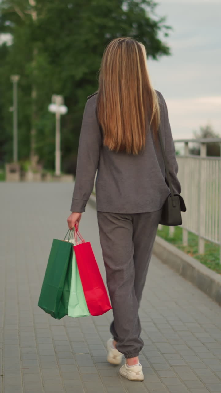 vista trasera de una dama con ropa gris caminando a lo largo de un carril de hierro con bolsas de compras y bolso de mano negro, con vegetación borrosa en el fondo, escena al aire libre casual durante un tranquilo paseo nocturno