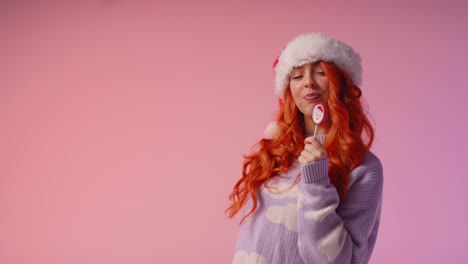 studio portrait shot of young gen z woman wearing christmas santa hat holding candy lollipop 1