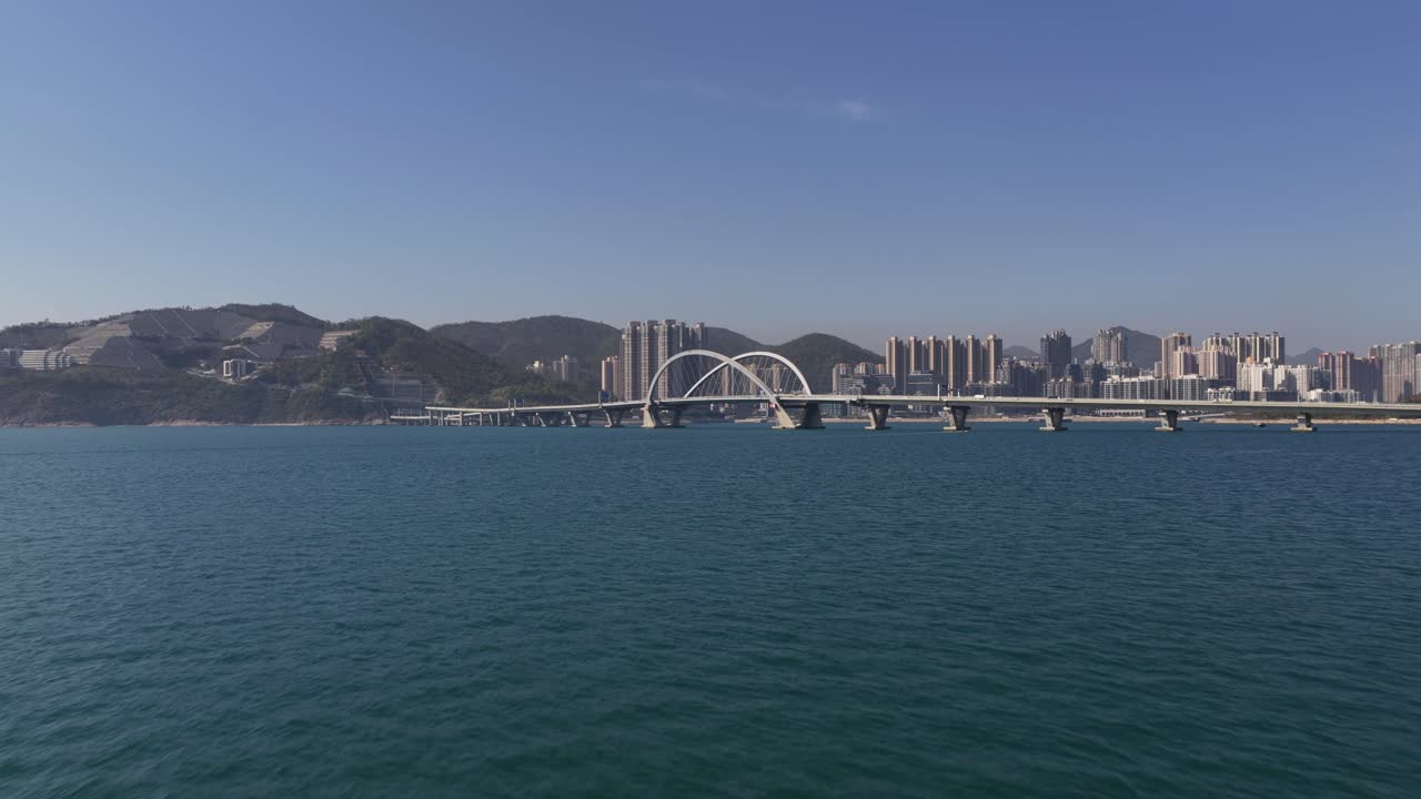 Splendida ripresa soggettiva da una barca che si avvicina a una moderna città costiera con un suggestivo ponte ad arco bianco, navigando su acque calme e turchesi sotto un cielo azzurro e limpido.