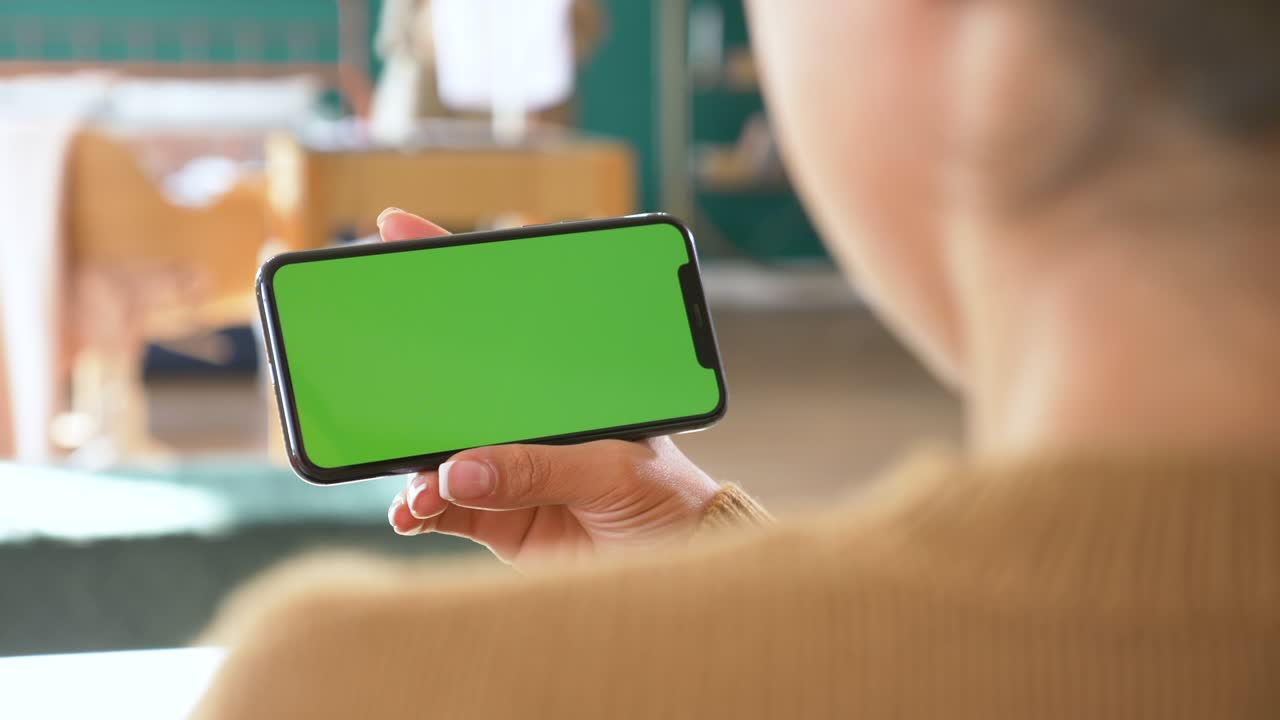 소파에서 휴식을 취하고 손을 흔들면서 수평으로 스마트폰 녹색 화면을 들고 있는 젊은 여성.