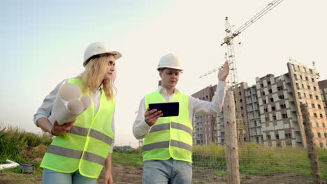 adult engineer man and architect woman uses a tablet in operation. writes a message or checks a drawing. against background is building. see neighborhood and drawings for verification of completed and planned construction works. the work of a crane