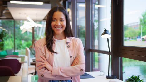 Portrait-Of-Smiling-Businesswoman-Standing-By-Desk-In-Modern-Open-Plan-Office