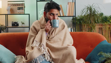 Sick-indian-man-wrapped-in-plaid-shivering-from-cold-drinking-hot-tea-in-unheated-apartment-due-debt