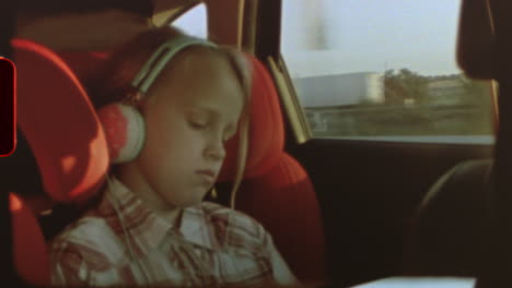 filmed on super 8 film blonde young girl peacefully resting in car seat, listening to music through headphones during family road trip, countryside landscape blurring outside window