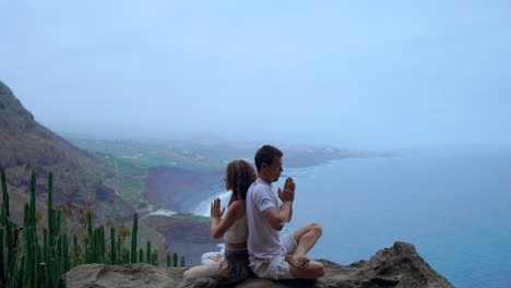 amidst mountainous surroundings and the ocean's beauty, a man and woman sit on a rock back to back, engaging in meditation and yoga