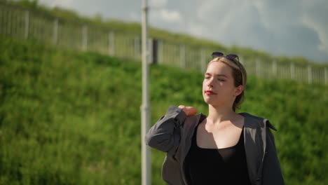 woman walking near hill with iron rails and greenery, carrying shopping bags backwards on her shoulder, wearing open shirt that reveals a black top, background of lush green hillside