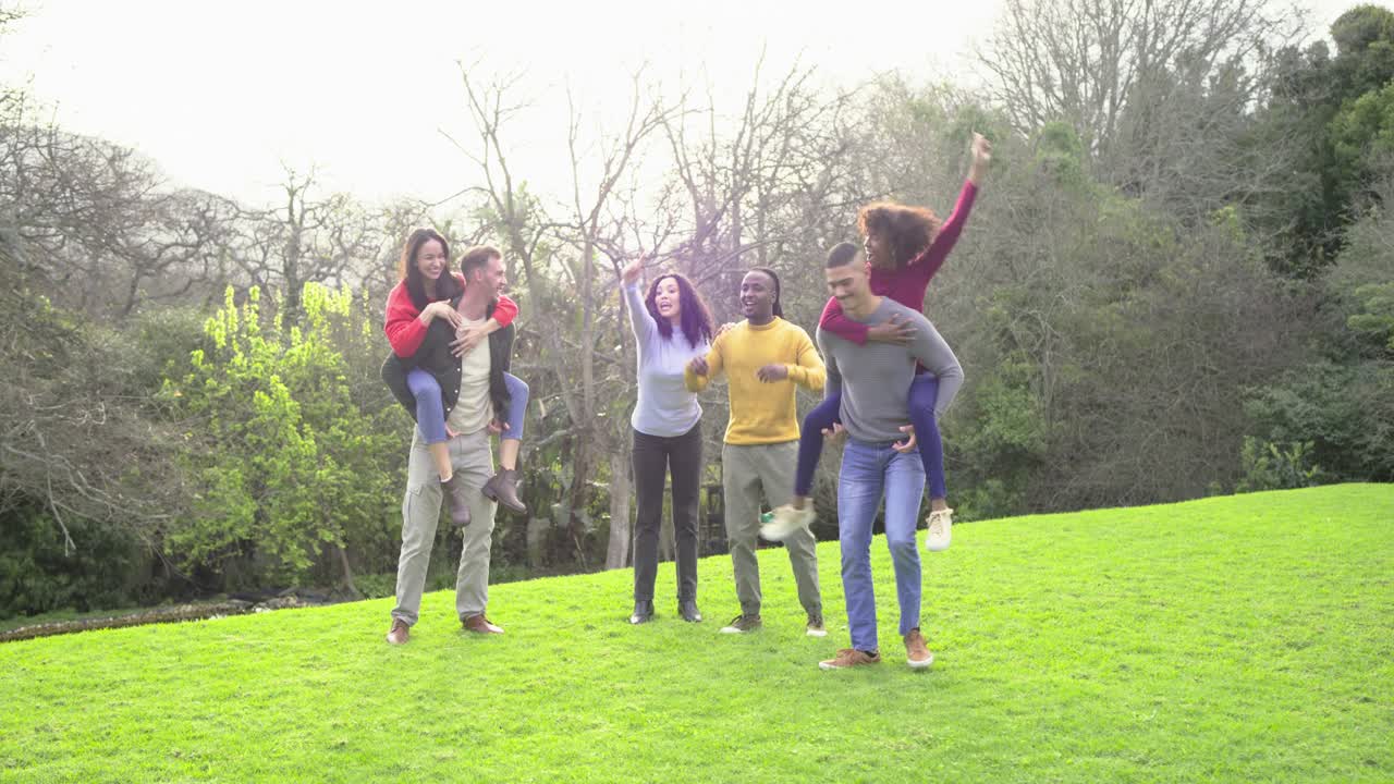 Diverse friends laughing, squatting then piggybacking partners and running on grassy slope