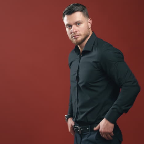 confident businessman in stylish wear. handsome man with beard posing on camera in dark red studio. portrait of young brunette male in black shirt.