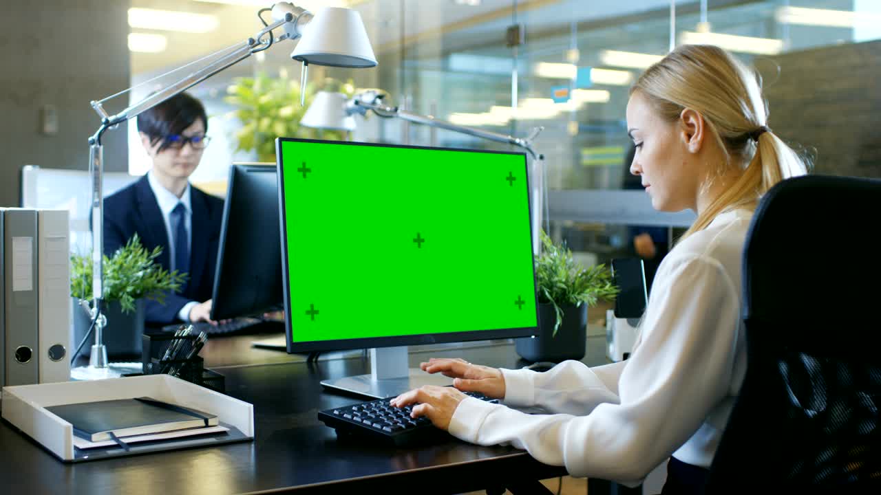 In the Office Businesswoman works at Her Desk on a Personal Computer with Mock-up Green Screen. Colleague Enters Office and Takes Place at His Desk.