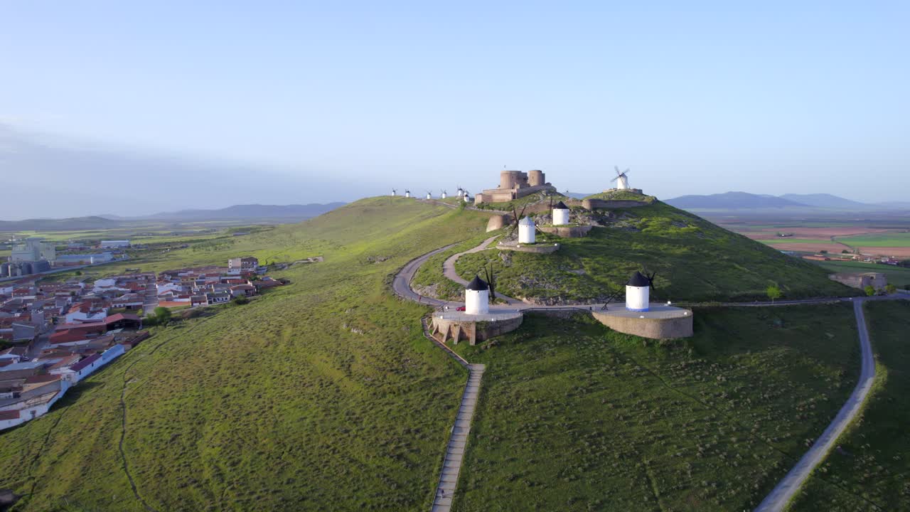 ascenso de drones que revela un conjunto de molinos de viento antiguos y un castillo medieval en la cima de una colina al amanecer