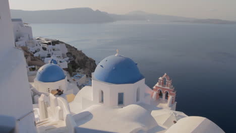 Cúpulas-De-Iglesia-Azul-Y-Campana-De-Iglesia-Rosa-Con-Vistas-A-Un-Majestuoso-Paisaje-Marino-En-Oia,-Santorini