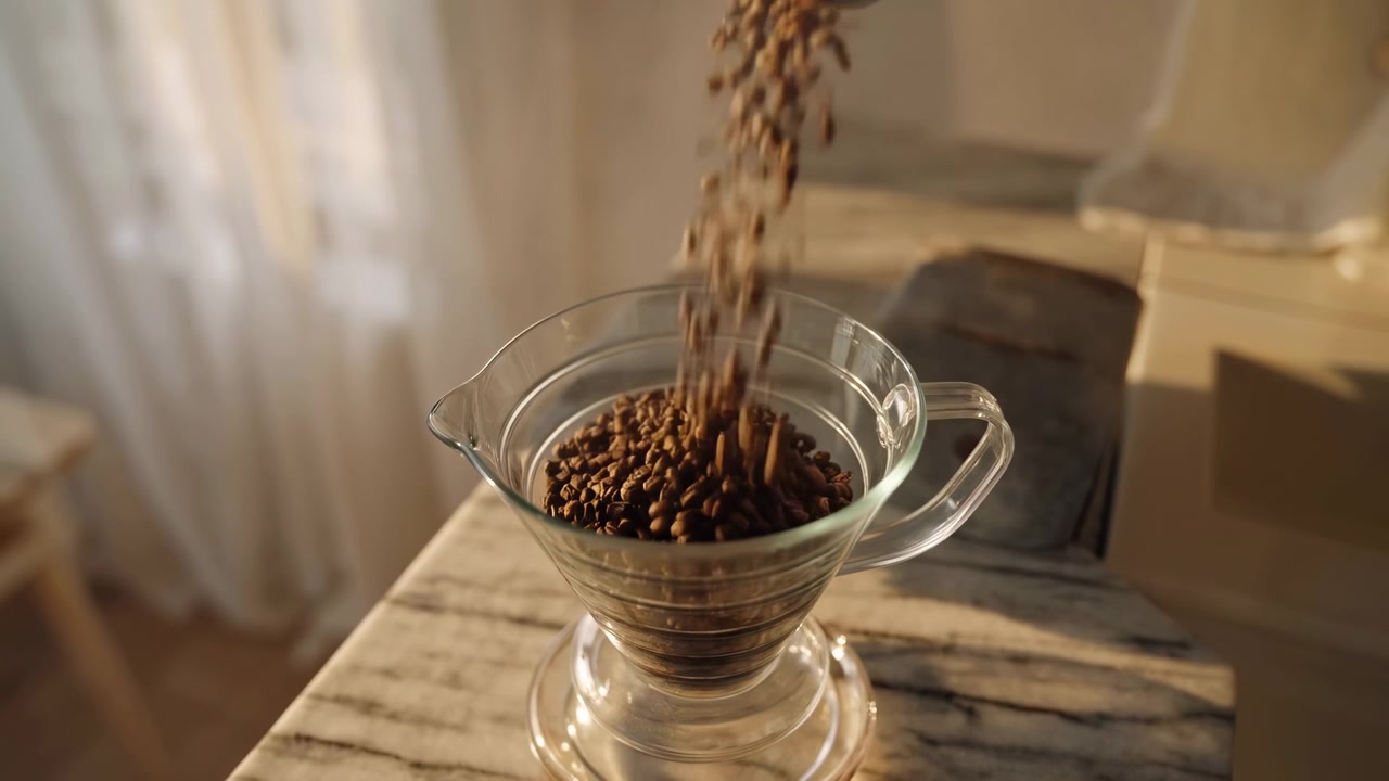 Friske kaffebønner falder ned i en klar glasdrypper på et rustikt træbord, oplyst af varmt indendørs lys med en blødt sløret baggrund og en hyggelig tilberedningsatmosfære.