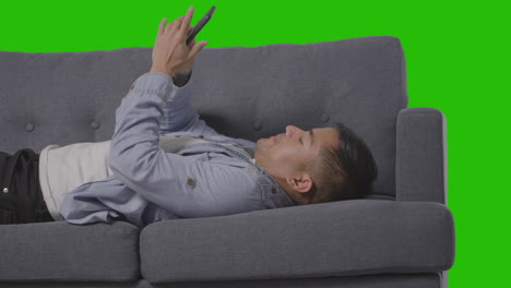 Studio-Shot-Of-Man-Lying-On-Sofa-Scrolling-Through-Mobile-Phone-And-Sneezing-Against-Green-Screen