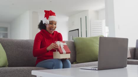 mujer con sombrero de santa abriendo caja de regalos mientras tiene video chat en su computadora portátil