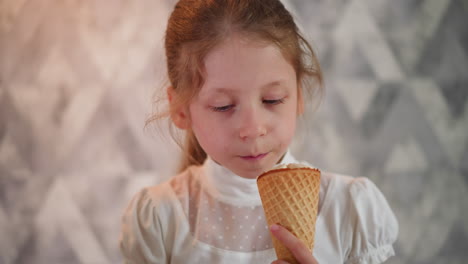 young girl in white top enjoying ice cream cone, gently licking it with eyes closed in relaxed delight, standing in front of gray geometric background