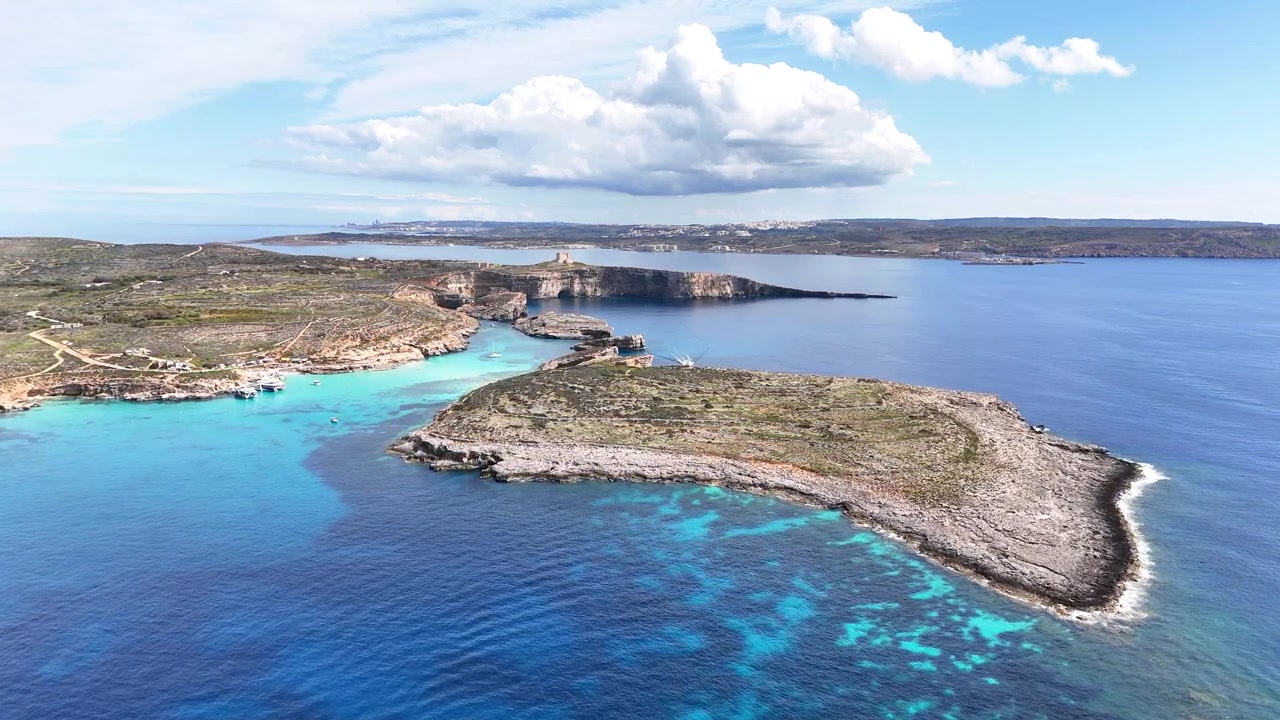 Laguna Biru antara Comino dan Cominotto Kemmunett dengan tebing Batu Kapur Koral Atas dan Menara St Mary Torri ta Santa Marija menghadap Laut Mediterania, bidikan udara lebar dari ketinggian