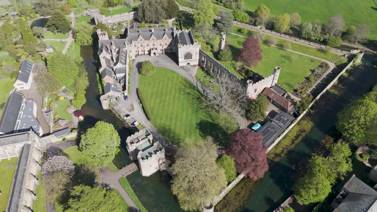 Aerial glide over moated medieval palace with crenellated walls, tranquil lawns and dense trees beside Wells cathedral precinct, portraying heritage serenity